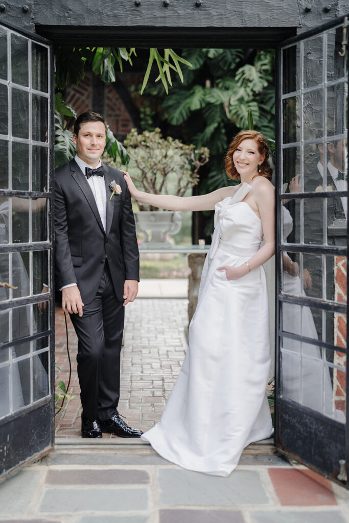 Bride and groom posing for the camera as the lean against glass doors, shot by natural light wedding photographers For Fearless Hearts