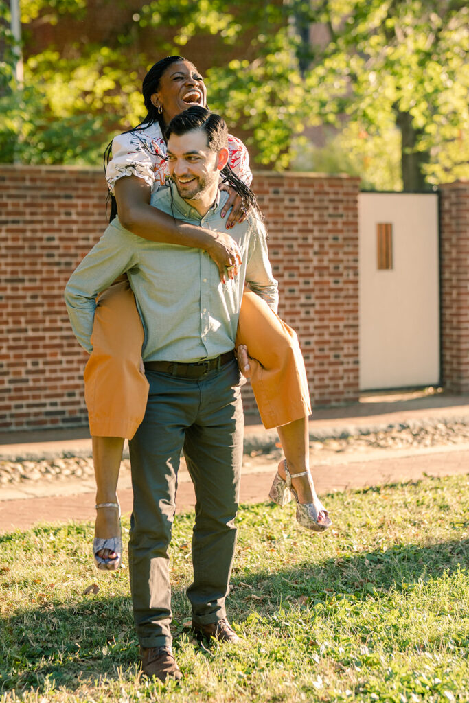 Guy smiling as he gives her fiance a piggyback ride during their engagement session