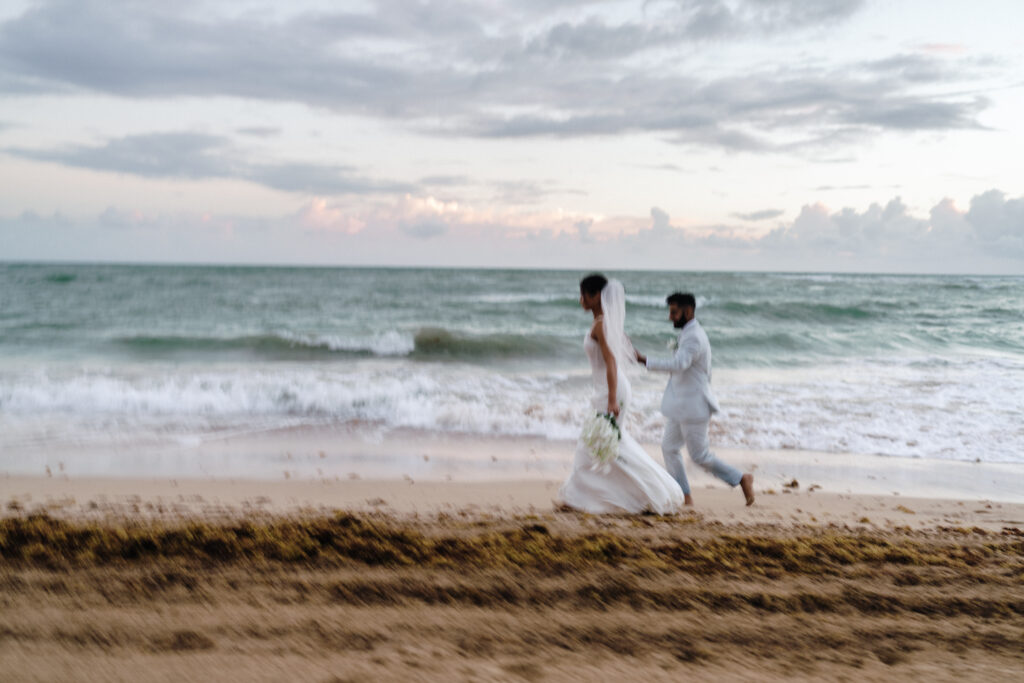 Blurry motion shot of newlywed couple running along the beach, captured by For Fearless Hearts